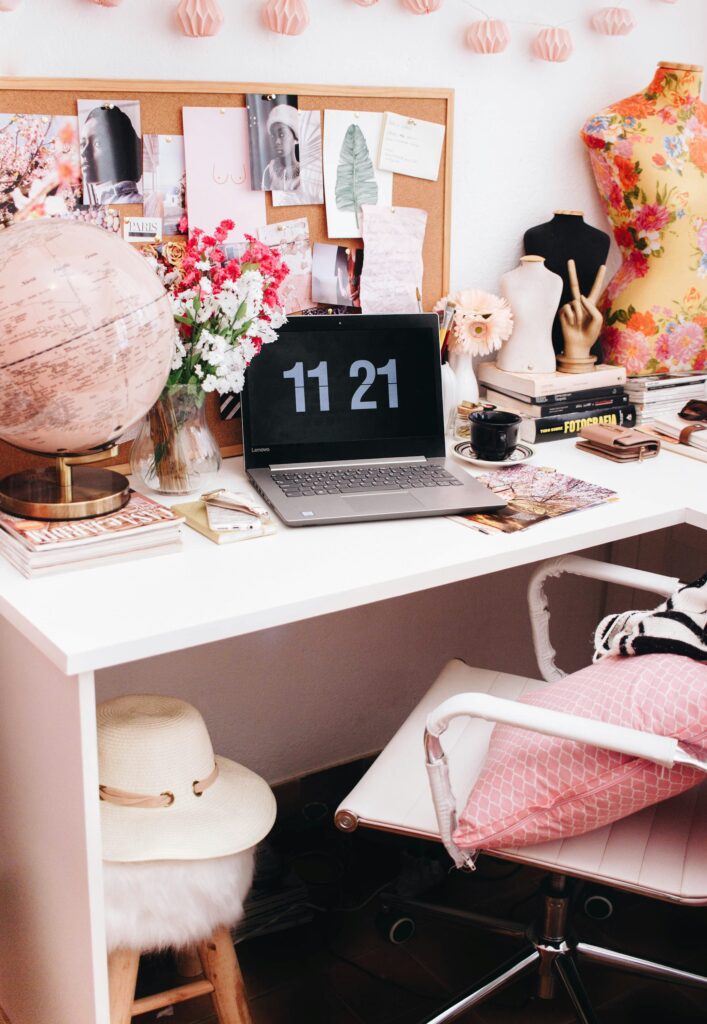 Pink aesthetic desk set up with laptop open stating the time as 11:21 in white letters against black background.  Pink and gold world globe and pinboard of eclectic postcards also appear on the desk.
